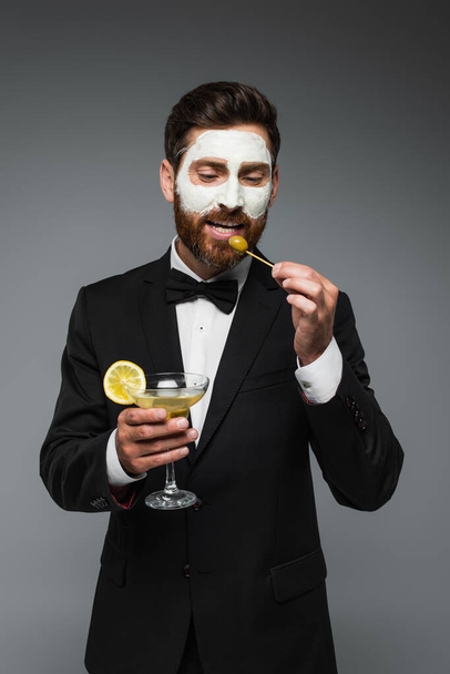 бородатый мужчина в костюме с глиняной маской на лице, держа коктейль и зубочистку с оливкой, изолированной на сером  - Фото, изображение
