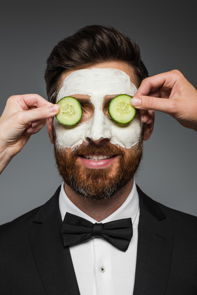 women applying sliced cucumbers on eyes of bearded man in suit with clay mask  isolated on grey  - 写真・画像