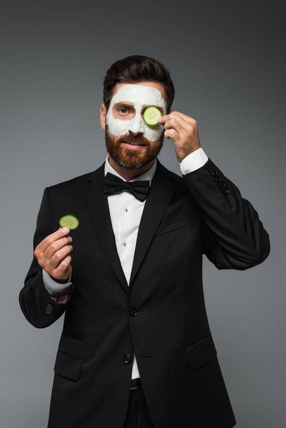 bearded man in suit with clay mask on face holding slices of cucumber isolated on grey  - Foto, Imagem