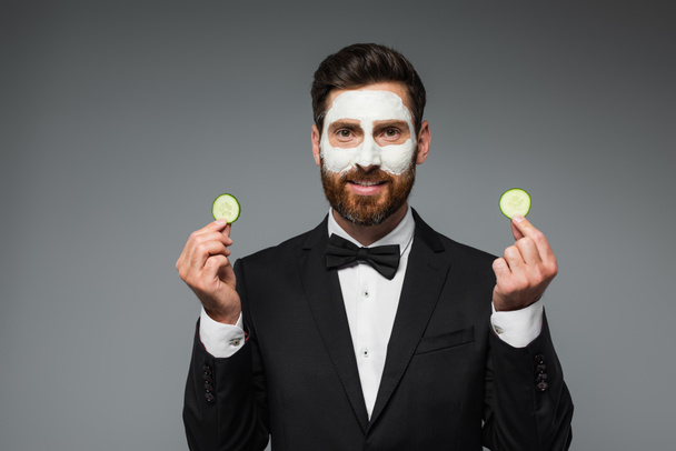 happy bearded man in suit with clay mask on face holding slices of cucumber isolated on grey  - Fotografie, Obrázek