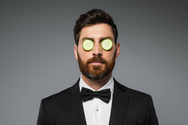 bearded man in tuxedo with fresh sliced cucumber on eyes isolated on grey - 写真・画像