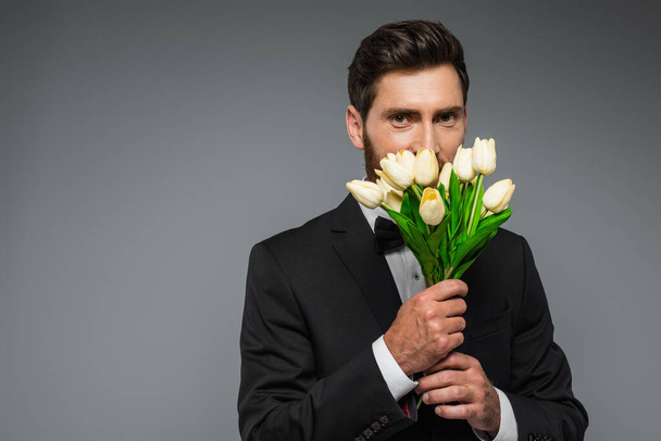 bearded man in elegant tuxedo smelling fresh tulips isolated on grey - 写真・画像