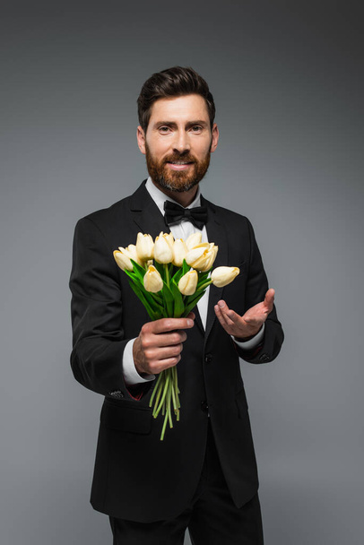 bearded man in elegant suit with bow tie holding fresh tulips and smiling isolated on grey  - Photo, Image