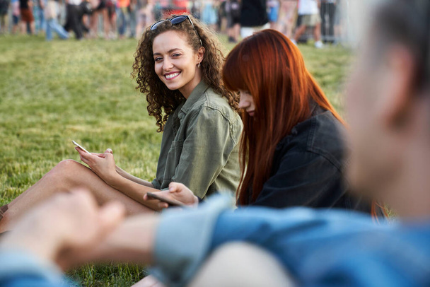 Γυναίκα κάθεται στο γρασίδι και χρησιμοποιώντας το κινητό τηλέφωνο στο φεστιβάλ με τους φίλους  - Φωτογραφία, εικόνα