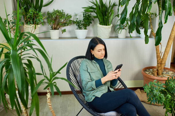 Woman hold mobile phone and surfing internet, type message, sitting on armchair at home garden, Biophilia design. - Fotoğraf, Görsel
