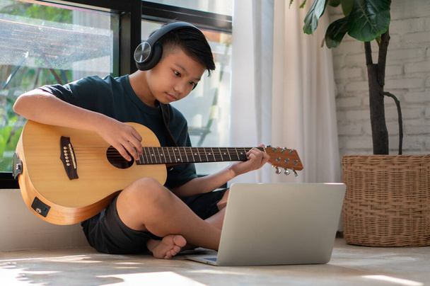 Ragazzo asiatico che suona la chitarra e guarda il corso online sul computer portatile mentre si esercita per imparare la musica o lo strumento musicale online a casa. Ragazzi studenti studiano online con gli insegnanti di videochiamata suonare la chitarra. - Foto, immagini