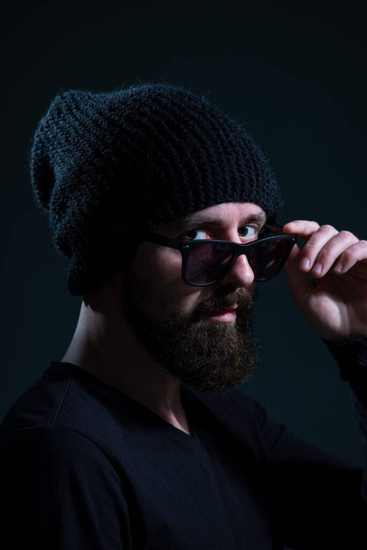authentic portrait of a bearded millennial in sunglasses and a black knitted hat against a dark background. - Photo, Image