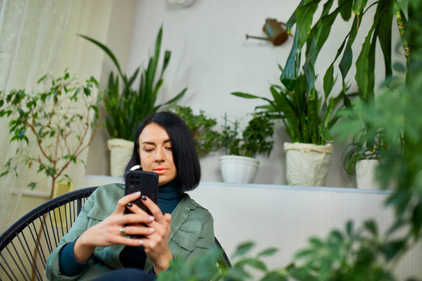 Женщина держит мобильный телефон и серфинг Интернет, тип сообщения, сидя на кресле в саду дома, дизайн Biophilia. - Фото, изображение