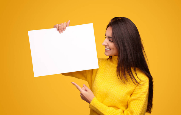 Cheerful young Hispanic brunette in sweater smiling and pointing at empty banner during advertisement campaign against yellow background - Photo, Image
