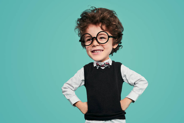 Cheerful funny little kid with dark wavy hair in classy clothes and round glasses, smiling brightly and looking away while standing against turquoise background with hands on waist - Photo, Image