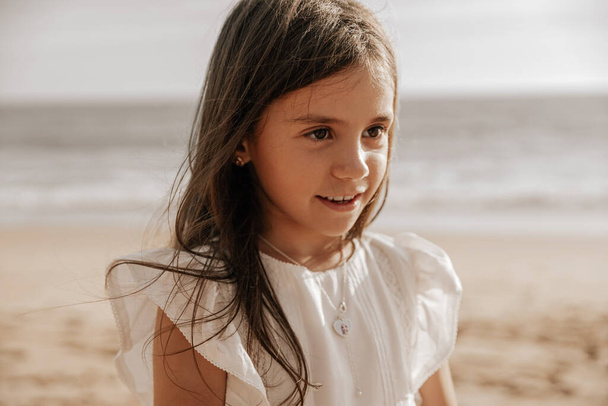 Cute child with long hair smiling and looking away while spending summer day on sandy beach near sea - Foto, immagini
