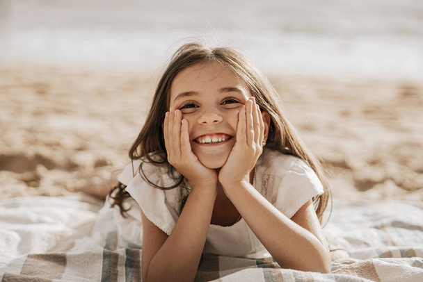 Happy girl kid lying with long hair smiling and looking at camera while lying on blanket on sandy beach - Fotó, kép