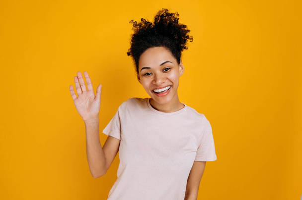 Greeting gesture. Positive pretty, curly haired african american young woman, waving hand, makes greeting gesture, looking at the camera, smiling, standing over isolated orange background - Photo, image
