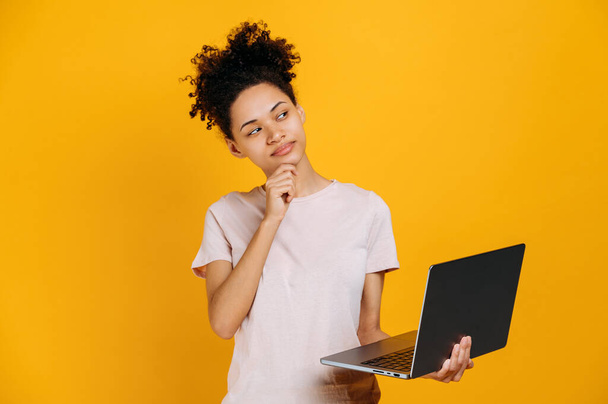 Thoughtful african american curly haired woman, looks away pensive, thinks about an idea, project, startup, holds an open laptop in her hand, stands on an isolated orange background, smiles, dreaming - Fotografie, Obrázek