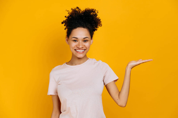 Photo of a friendly african american young woman in casual t-shirt, looks at the camera and points hand to the side, at empty space for presentation or text, stands on isolated blue background, smiles - Foto, immagini