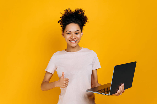 Positive lovely curly haired african american young woman, wearing casual clothes, holding an open laptop in her hand, looks at camera, showing thumbs up gesture, stands on isolated orange background - Фото, изображение