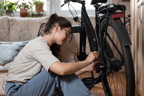 Young self learned woman or girl mechanic fixing and repairing her bicycle at her home. Female repair chain on rear wheel before she start to ride a bike on nice sunny weather. - Фото, изображение