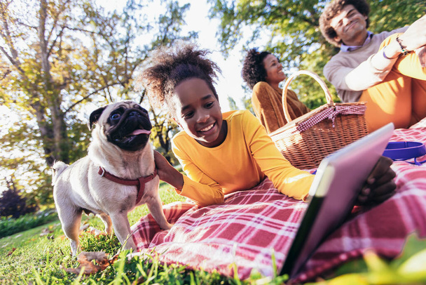 Beautiful young family lying on a picnic blanket with their dog, enjoying an autumn day in park while using digital tablet. - Foto, immagini