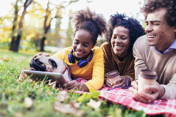 Beautiful young family lying on a picnic blanket with their dog, enjoying an autumn day in park while using digital tablet. - Photo, Image