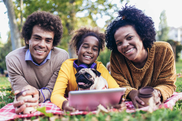 Beautiful young family lying on a picnic blanket with their dog, enjoying an autumn day in park while using digital tablet. - Valokuva, kuva