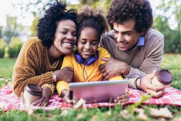 Hermosa familia joven acostada en una manta de picnic, disfrutando de un día de otoño en el parque mientras usa la tableta digital
. - Foto, Imagen