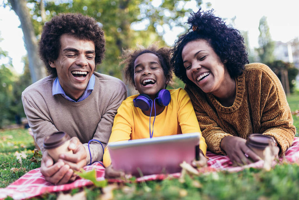 Schöne junge Familie liegt auf einer Picknickdecke und genießt einen Herbsttag im Park, während sie das digitale Tablet benutzt. - Foto, Bild