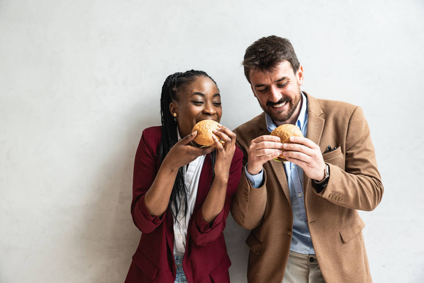 Two young business people or smart casual white man and African American woman holding and eating fat tasty American hamburger or burger while they making funny faces and goofing. Happy people eating  - Fotoğraf, Görsel