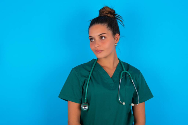 Mavi arka planda yeşil üniforma giyen güzel İspanyol doktor kadın boş alana dikkatlice bakıyor. - Fotoğraf, Görsel