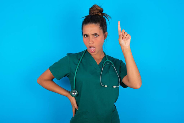 beautiful hispanic doctor woman wearing green medical uniform over blue background holding finger up having idea and posing - Photo, Image