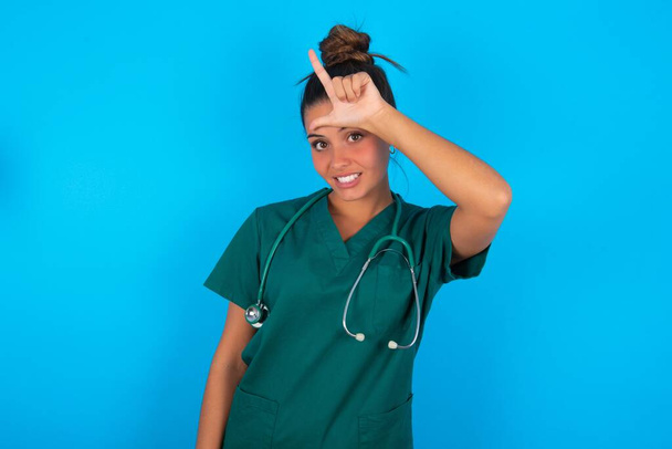 bela mulher médica hispânica vestindo uniforme médico verde sobre fundo azul zombando de pessoas com dedos na testa fazendo gesto perdedor zombando e insultando. - Foto, Imagem