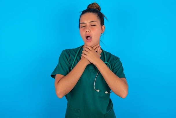 schöne hispanische Ärztin in grüner Arztuniform vor blauem Hintergrund schreit ersticken, weil schmerzhafte Würgen. Gesundheitsprobleme. Asphyxid- und Selbstmordkonzept. - Foto, Bild