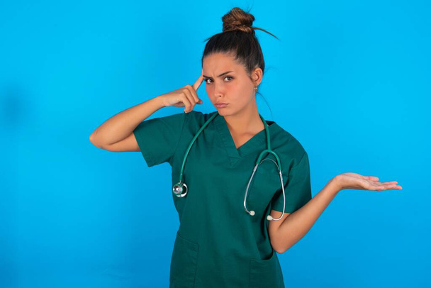bella donna medico ispanico indossa uniforme medica verde su sfondo blu confuso e infastidito con palmo aperto mostrando spazio copia e puntando il dito sulla fronte. Pensaci.. - Foto, immagini