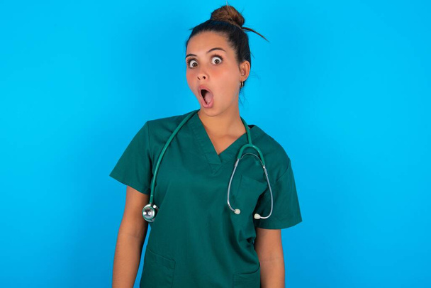 Oh, meu Deus. Surpreendida bela mulher médica hispânica vestindo uniforme médico verde sobre fundo azul olha para a câmera com expressão chocada exclama com inesperado, - Foto, Imagem