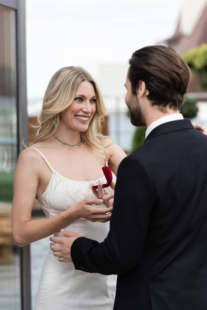 Young man holding box with engagement ring near smiling girlfriend on terrace  - Foto, Bild