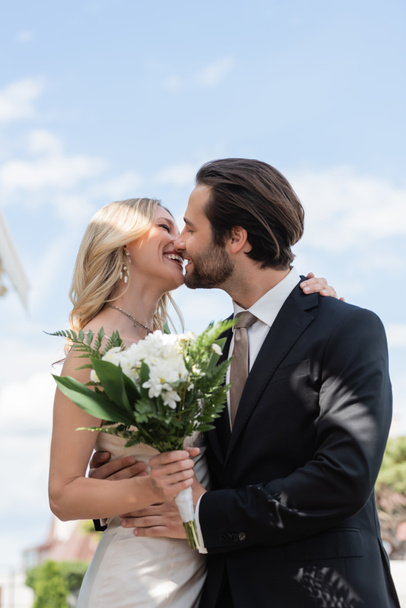 Side view of cheerful newlyweds holding flowers and kissing outdoors  - Foto, Imagem