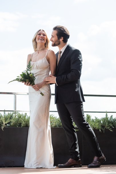 Positive groom in suit hugging bride in dress holding bouquet on terrace of restaurant  - 写真・画像