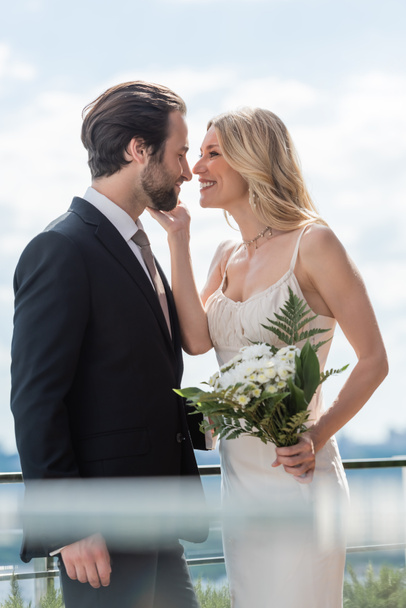 Side view of smiling bride with bouquet touching face of bearded groom on terrace  - Photo, image