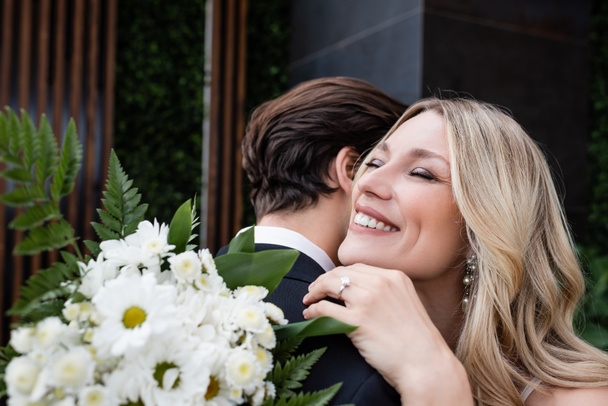 Cheerful bride embracing groom near bouquet outdoors  - Zdjęcie, obraz