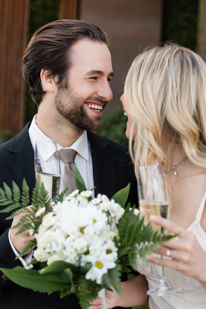 Positive groom holding glass of champagne near bride with bouquet outdoors  - Foto, Bild