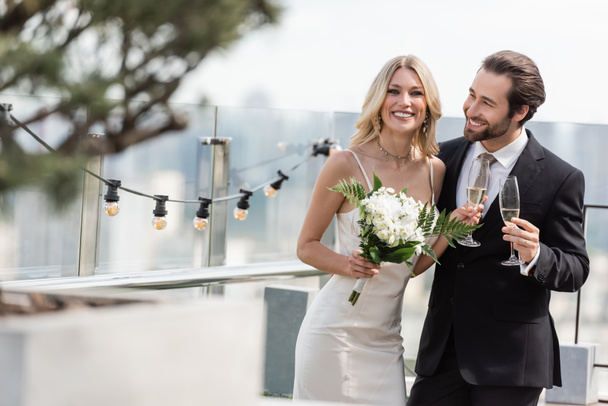 Smiling bride with bouquet and champagne looking at camera near elegant groom on terrace  - Foto, imagen