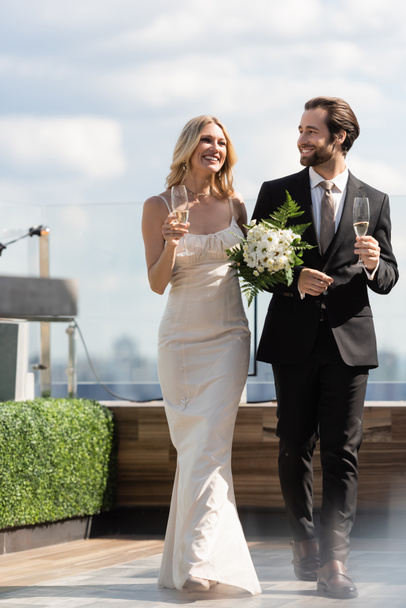 Positive newlyweds with flowers and champagne walking on terrace of restaurant  - Foto, Bild