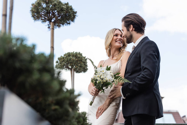 Low angle view of groom looking at smiling bride with flowers on terrace  - Fotoğraf, Görsel