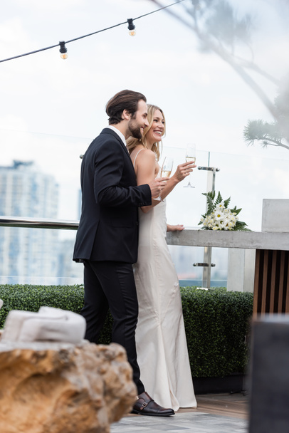 Smiling groom in suit holding glass of champagne near bride and bouquet on terrace  - 写真・画像