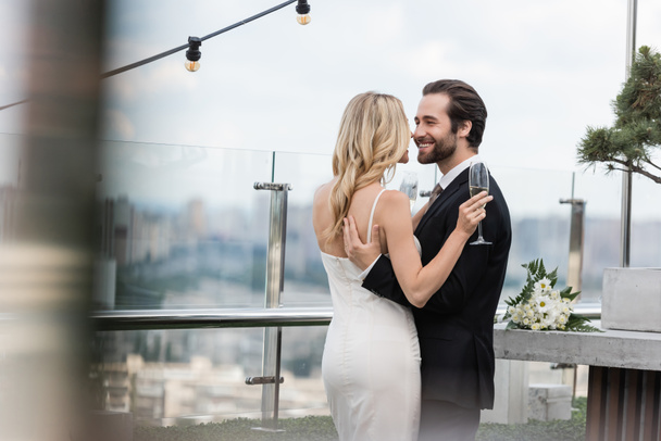 Positive groom hugging bride with glass of champagne on terrace of restaurant  - Valokuva, kuva