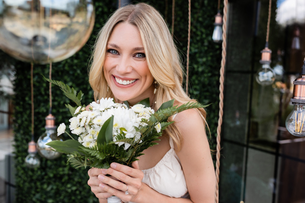 Portrait of positive bride holding bouquet and looking at camera near blurred light bulbs outdoors  - Foto, Imagem