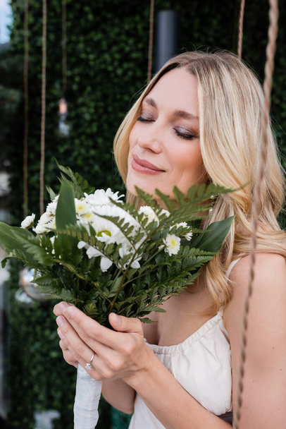 Blonde bride with closed eyes smelling bouquet on terrace  - Фото, изображение