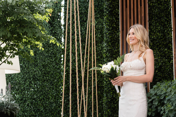 Positive bride holding flowers near plants on terrace  - Foto, afbeelding
