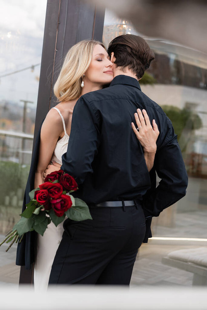 Blonde woman in dress holding red roses and hugging boyfriend outdoors  - Fotó, kép