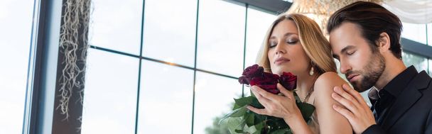 Brunette man in suit touching shoulder of blonde girlfriend with roses in restaurant, banner  - Foto, afbeelding
