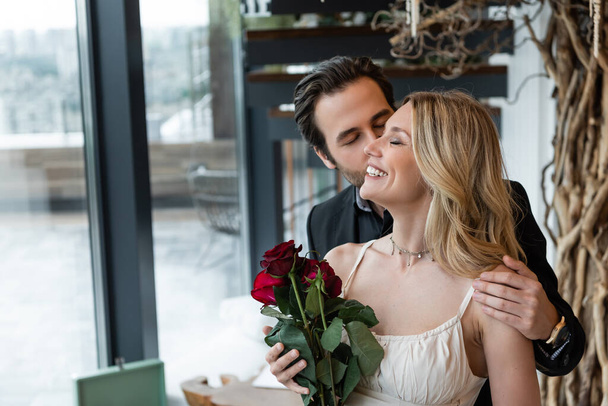 Brunette man kissing cheerful girlfriend with red roses in restaurant  - 写真・画像